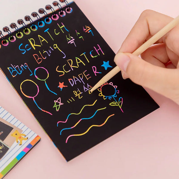 A hand is drawing on a black notepad with colorful markers, creating scratch art designs.