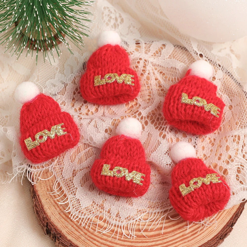 A set of mini knitted red hats with white pom-poms and gold LOVE embroidery displayed on a lace fabric background.