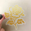 A transparent sticker featuring a gold-colored rose and leaf botanical design on a white background.