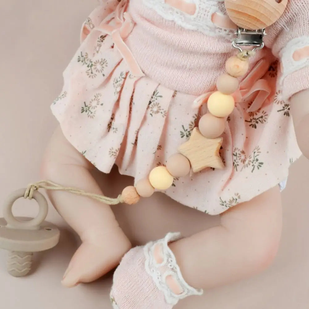A baby wearing a pink ruffled top and a wooden pacifier clip with silicone beads on a string.