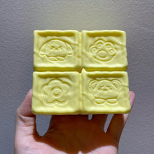 A hand holds a block of yellow strawberry milk squishy stress relief toy with various molded shapes.