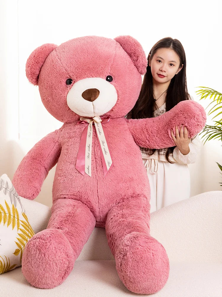 A large pink teddy bear plush toy sitting on a white sofa next to a young woman.