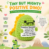 A handmade crochet dinosaur doll in green and yellow with a motivational card encouraging courage.