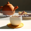 A white cup filled with brown liquid sits on an orange saucer, with a terracotta teapot and waffle cookies in the background.