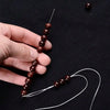 A hand holds a string of dark brown metal beads threaded onto a needle, with loose threads extending from the beads.