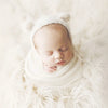 A newborn baby is sleeping wearing a white knit bear hat and wrap set surrounded by fluffy white material.