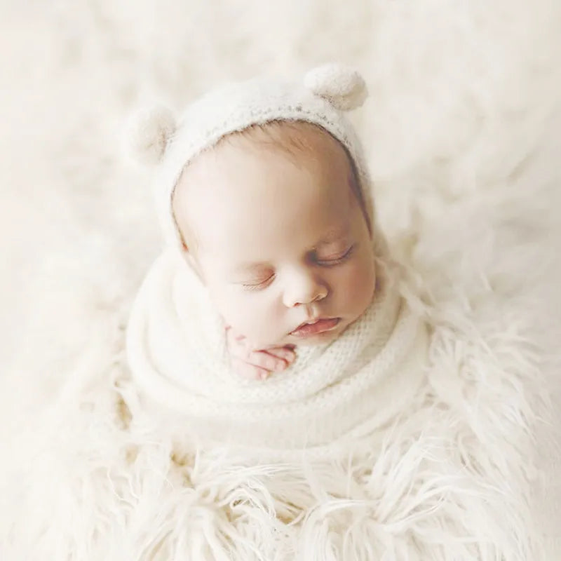 A newborn baby is sleeping wearing a white knit bear hat and wrap set surrounded by fluffy white material.