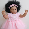 A lifelike reborn toddler doll with hand-rooted black hair and a pink lace dress.