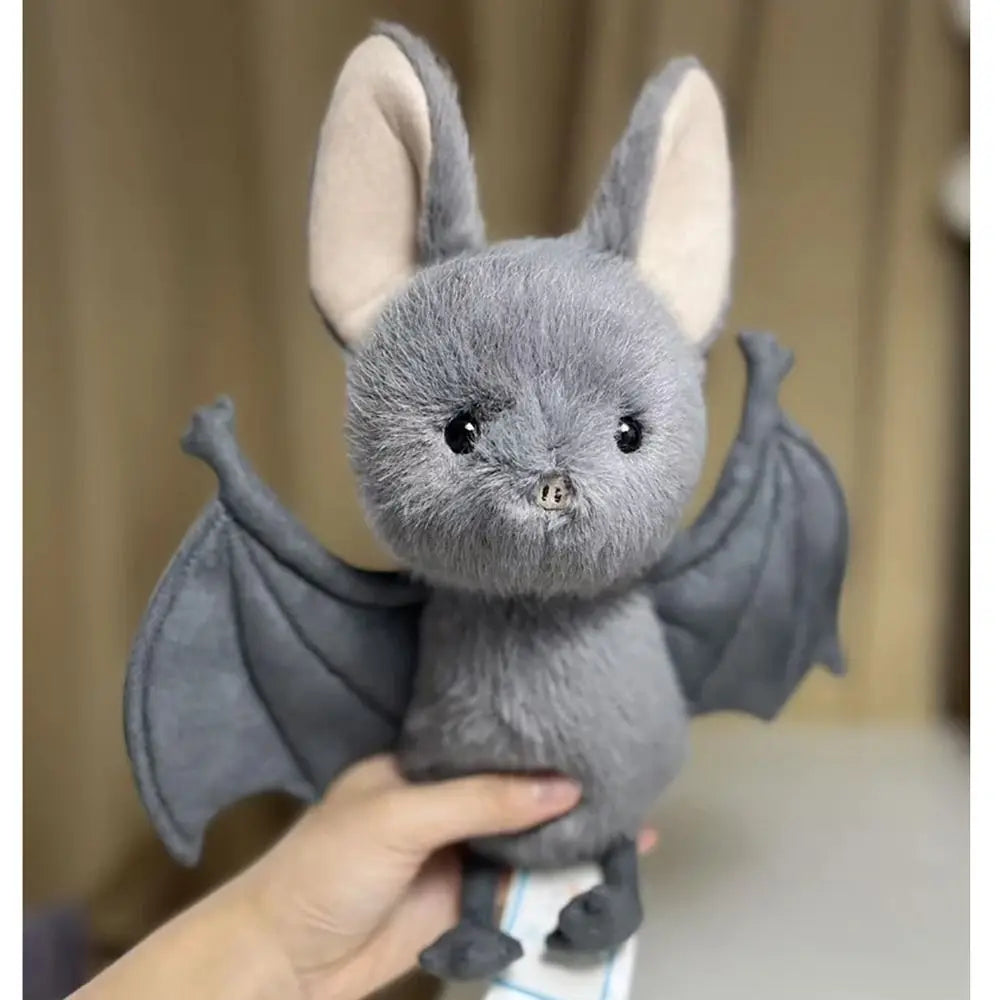 A grey bat plush toy with large ears and wings is being held by a hand.