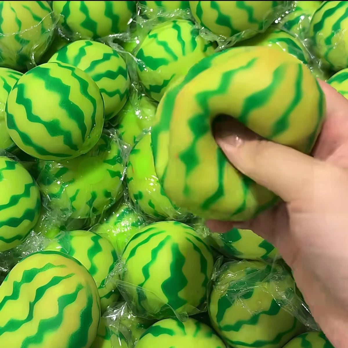 A hand is squeezing a bright green watermelon-themed squishy stress ball with a striped texture.