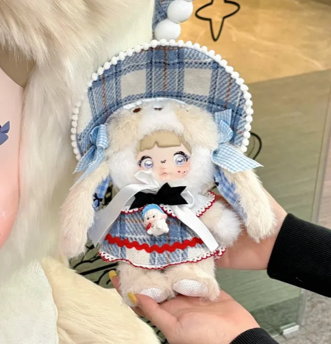 A fluffy white doll wearing a blue plaid bonnet and dress is held in a person's hand.