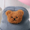A knitted baby hat shaped like a brown teddy bear is shown against a light blue background.