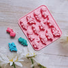 A pink silicone mold with dinosaur shapes is displayed on a white wooden surface surrounded by white flowers.