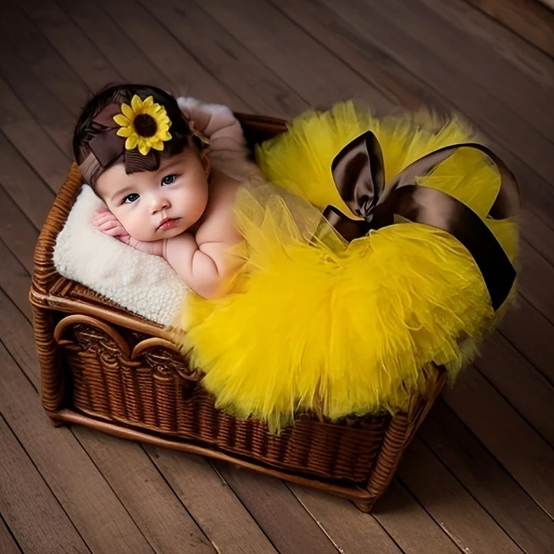 A baby wearing a yellow tutu skirt and headdress set with a sunflower decoration, resting in a woven basket.