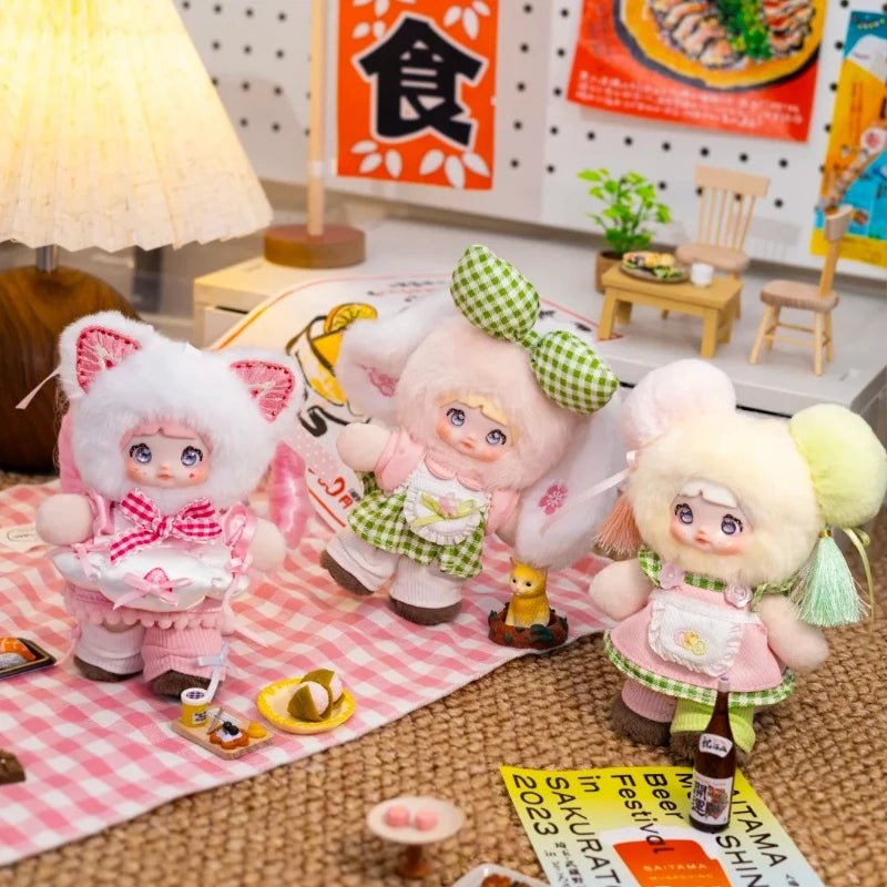 A plush keychain of a pink animal character with green accents displayed on a pink checkered cloth.