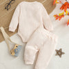 A light pink knitted baby romper and pants set lying on a beige woven fabric with some autumn leaves.