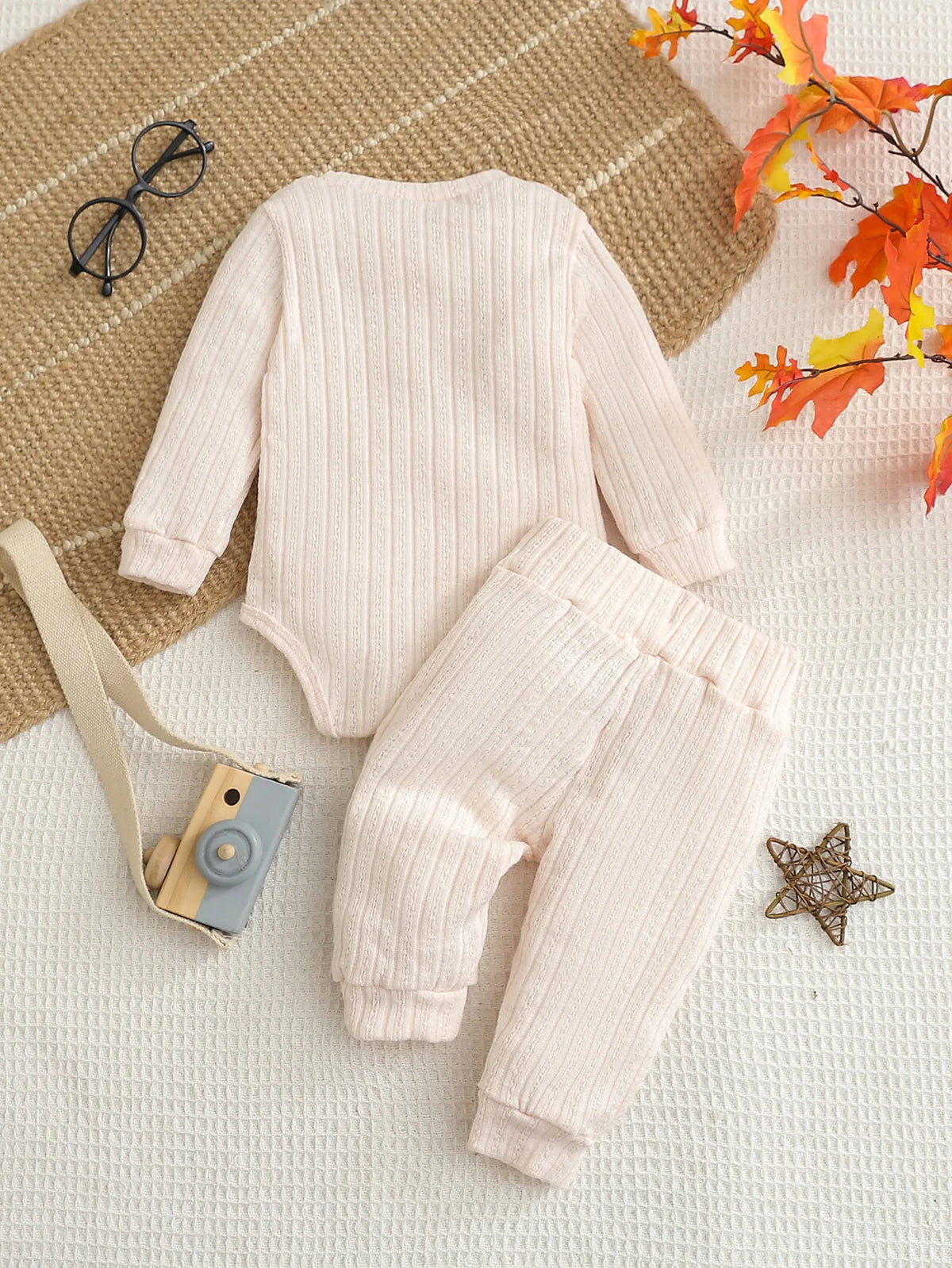 A light pink knitted baby romper and pants set lying on a beige woven fabric with some autumn leaves.