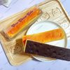 A slice of orange cheese cake and a dark chocolate butter stick squishy toy are displayed on a wooden cutting board.