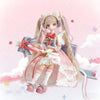 A female doll with long blonde hair and pink clothing wearing a bow and wings, displayed in a blind box packaging.