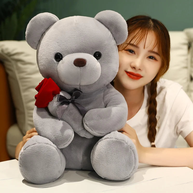 A woman is posing with a gray teddy bear plush toy that has a red rose attached.