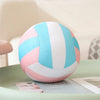 A soft plush volleyball pillow with pink and light blue stripes resting on a green surface.