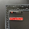 A close-up of a silver umbrella corporation car emblem badge and a red and white striped badge against a ruler.