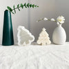 A set of white silicone Christmas cookie and candle molds displayed on a white cloth with green foliage.