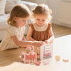 Two young girls play with a pink wooden rocket stacking toy and shape sorter on a light wood table.