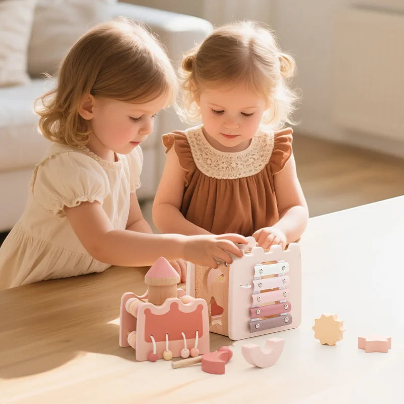 Two young girls play with a pink wooden rocket stacking toy and shape sorter on a light wood table.