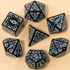 A set of seven fantasy carved polyhedral dice in black and white on a beige surface.
