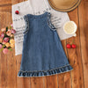 A denim sleeveless dress for a toddler girl with lace trim, resting on a wooden surface next to a straw hat and a cup.