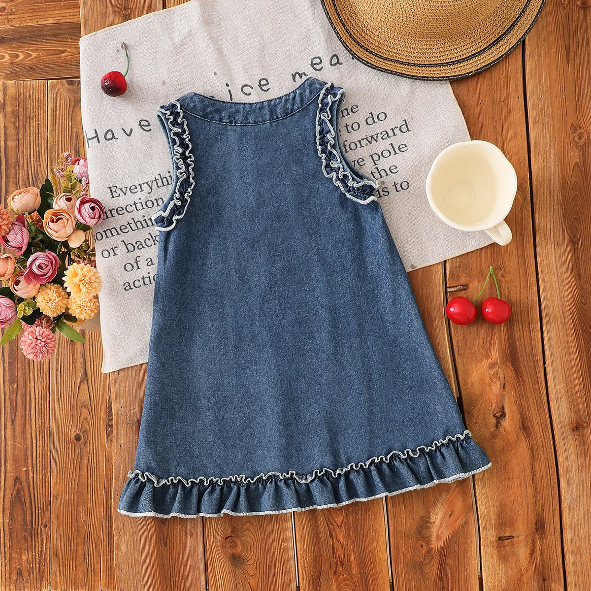 A denim sleeveless dress for a toddler girl with lace trim, resting on a wooden surface next to a straw hat and a cup.