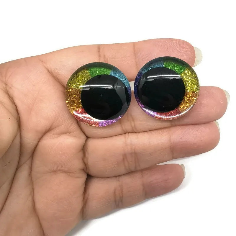 Pair of glitter cartoon safety eyes with black pupils and colorful irises displayed in a hand.
