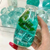 A hand holds a clear bottle filled with blue-green liquid and ice cubes, labeled as a chibi toy.