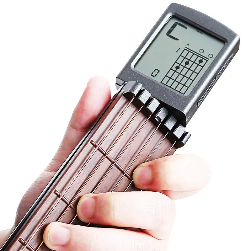 A hand holds a pocket guitar chord trainer with a wooden neck and a digital LCD display showing fret positions.