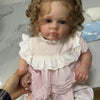 A lifelike reborn baby doll with pink dress and accessories is being held by a hand.