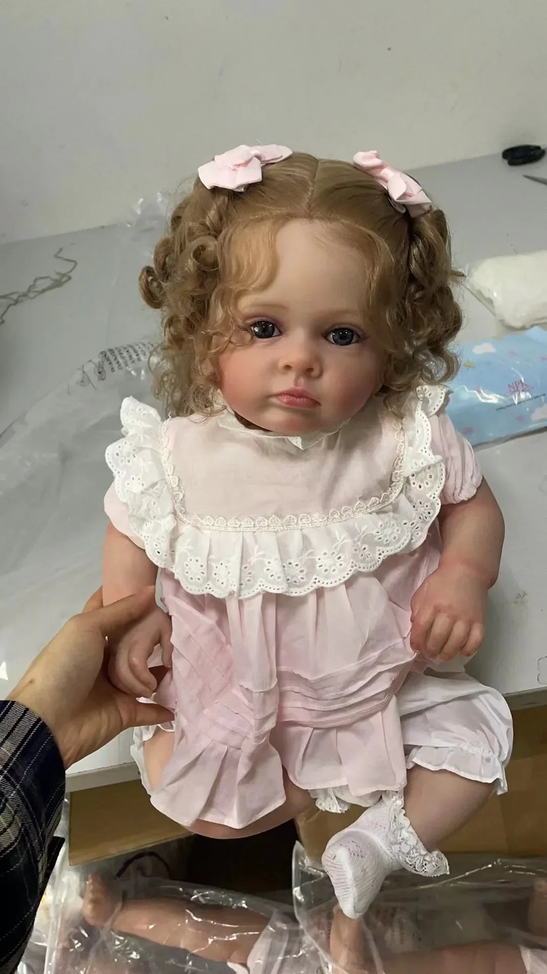 A lifelike reborn baby doll with pink dress and accessories is being held by a hand.