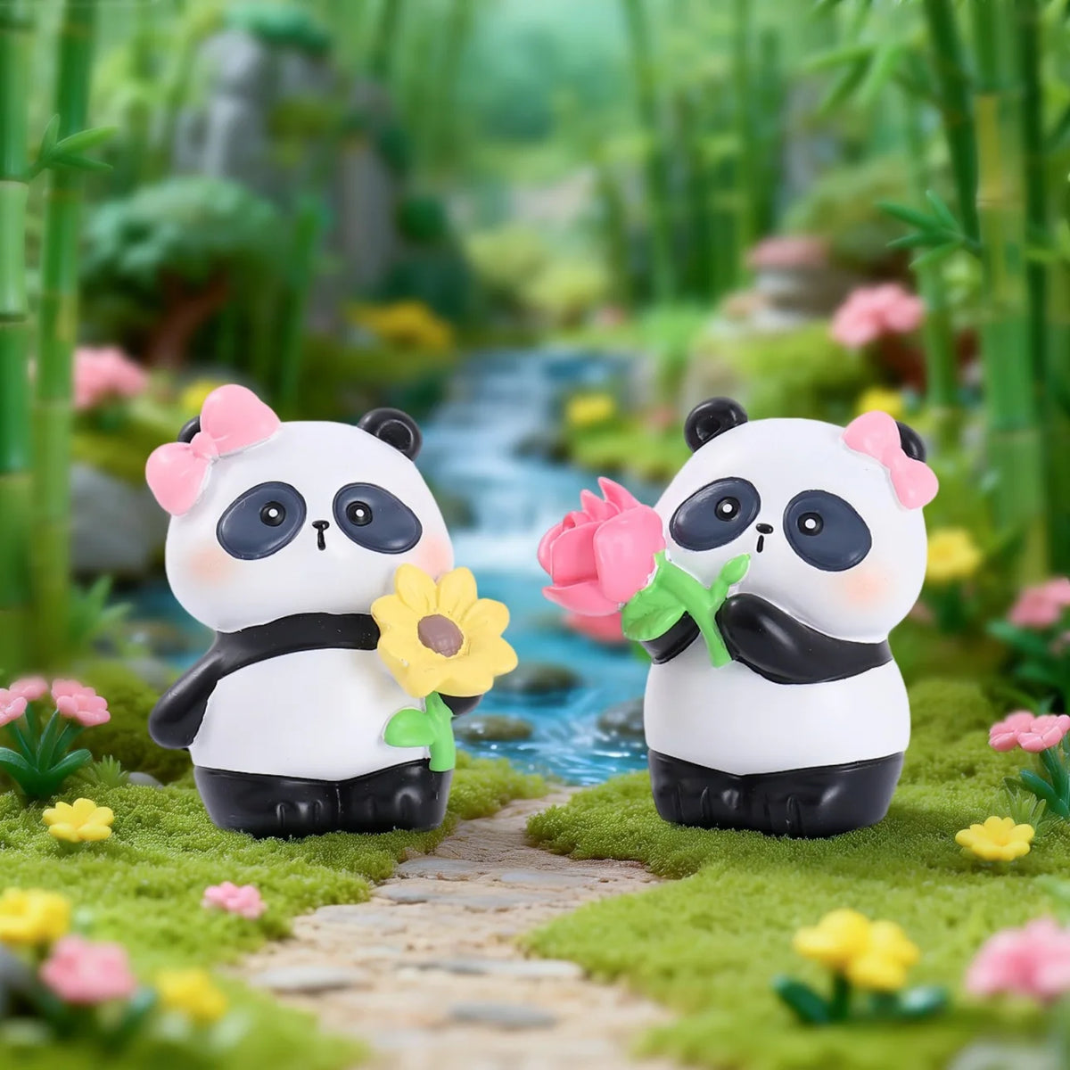 A pair of cute panda collectible figures in white and black sitting on a mossy path surrounded by green foliage.