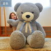 A gray giant plush teddy bear with a light yellow nose and blue ribbon tied around its neck sitting on a beige rug.