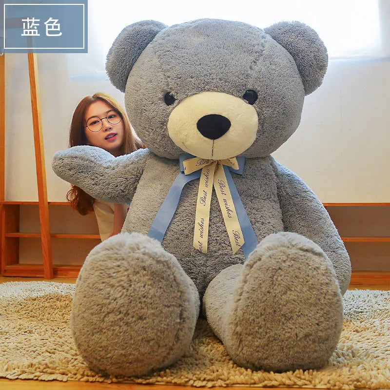 A gray giant plush teddy bear with a light yellow nose and blue ribbon tied around its neck sitting on a beige rug.