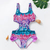 A vibrant pink and blue mermaid unicorn one-piece swimsuit lying on a white surface next to a green leaf and a seashell.