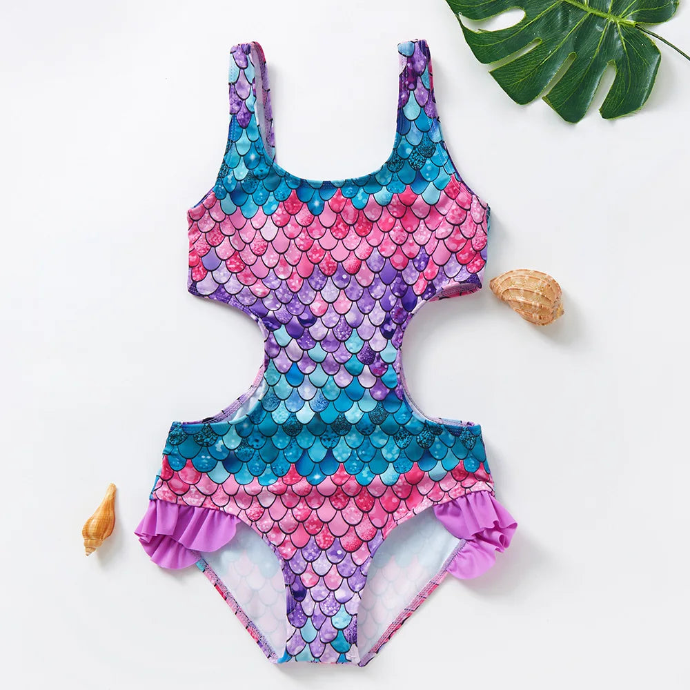 A vibrant pink and blue mermaid unicorn one-piece swimsuit lying on a white surface next to a green leaf and a seashell.