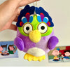 A purple and green plush toy of an owl with blue feathers and large eyes being held by a hand.