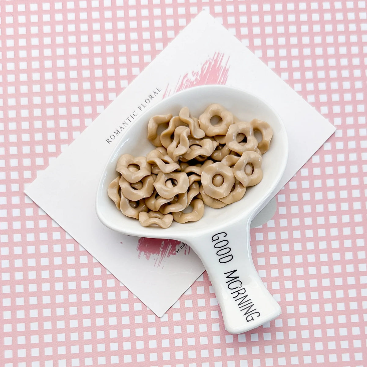 A white ceramic spoon holds light beige wavy acrylic beads with a floral edge on a pink patterned background.