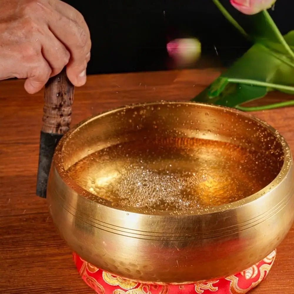 A golden Tibetan singing bowl resting on a red patterned cloth, with a hand holding a mallet nearby.
