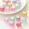 A collection of colorful floral square resin beads in pink, purple, and green are displayed on a white plate.