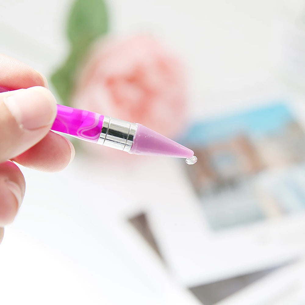 A hand holds a purple double-head diamond painting pen with a wax tip against a blurred background.