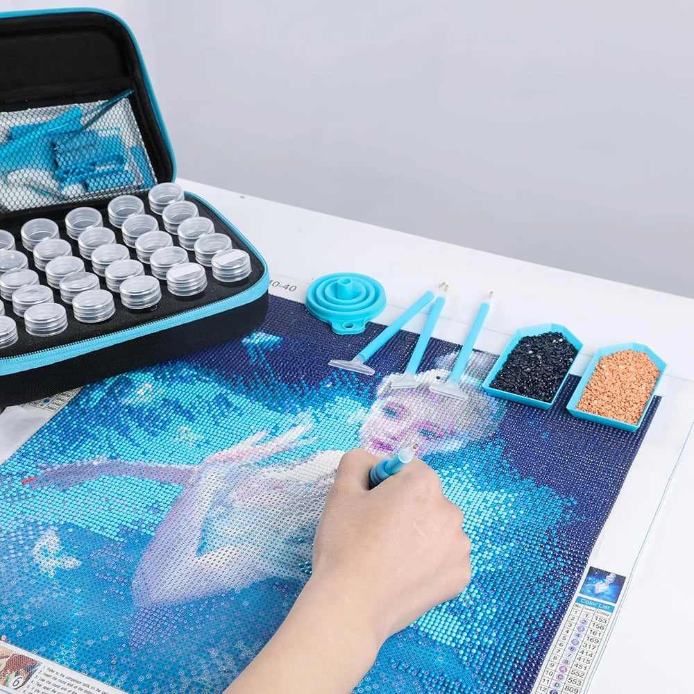 A hand is painting a diamond art piece on a blue background using a tool, with a storage case and jars visible.