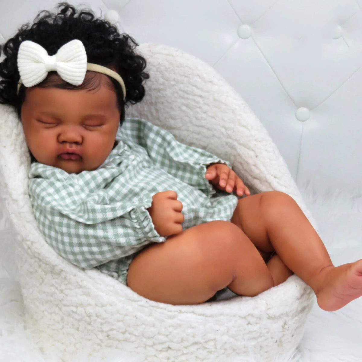 A newborn baby with dark curly hair wearing a green and white checkered outfit is resting in a white knitted basket.
