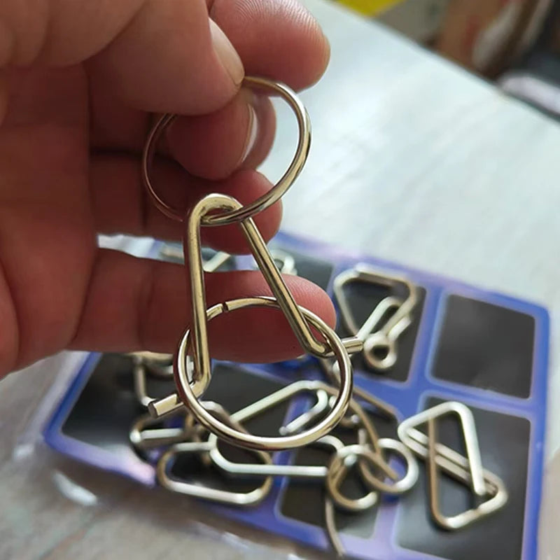 A hand holds a small metal wire puzzle chain with various interlocking rings and shapes.